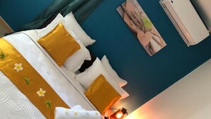 Een net opgemaakt bed met wit en geel beddengoed, een blauwgroene wand en een klein tafeltje met een lantaarn.