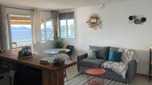 Een woonkamer met een grijze bank, een klein rond tafeltje en uitzicht op de zee door grote ramen.