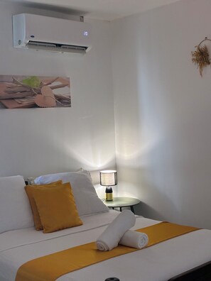 Hotellihuone, jossa on sänky, yöpöytälamppu, ilmastointilaite ja seinälle kiinnitetty taideteos.