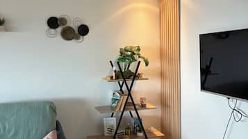 A modern living room with a sofa, a multi-tiered wooden shelf with decorative items, a wall-mounted clock, and a flat-screen TV.