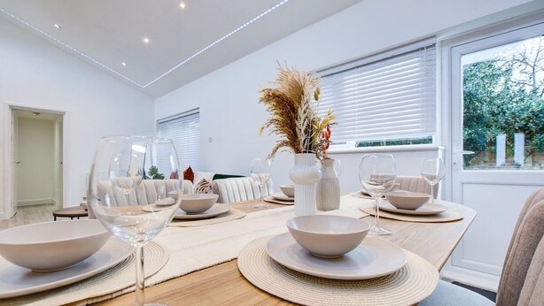 A modern dining area with a wooden table set for a meal, surrounded by white furniture and a large window with blinds. - Sleek & Exquisite 3 Bedroom Home (Colchester)