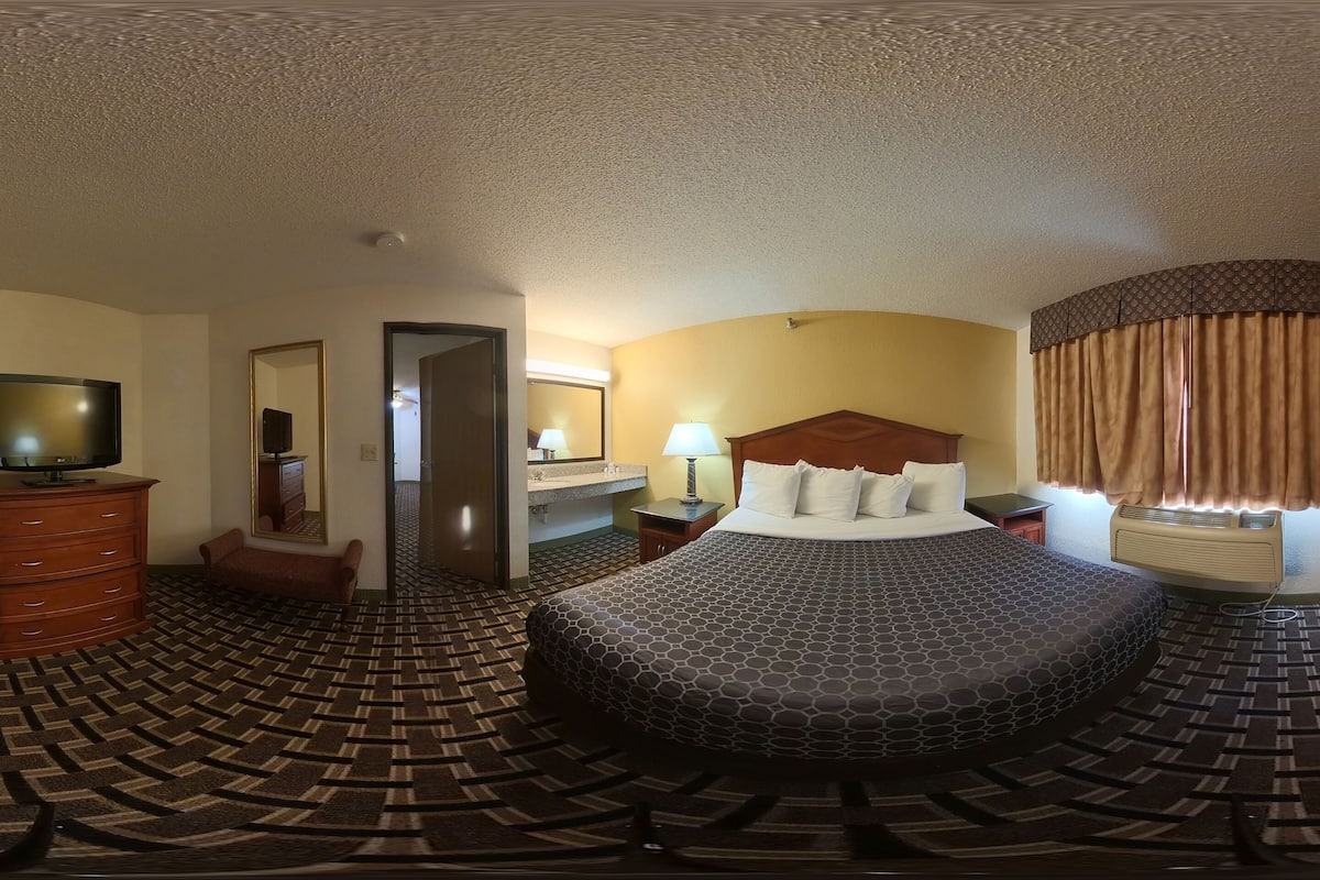 A hotel room with a large bed, a TV, a dresser, a mirror, a lamp, and a window with curtains.