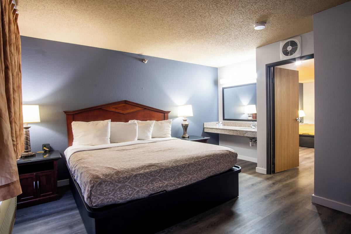 A hotel room with a large bed, a wooden headboard, two bedside tables with lamps, a nightstand, a bathroom with a mirror, and a door leading to another room.