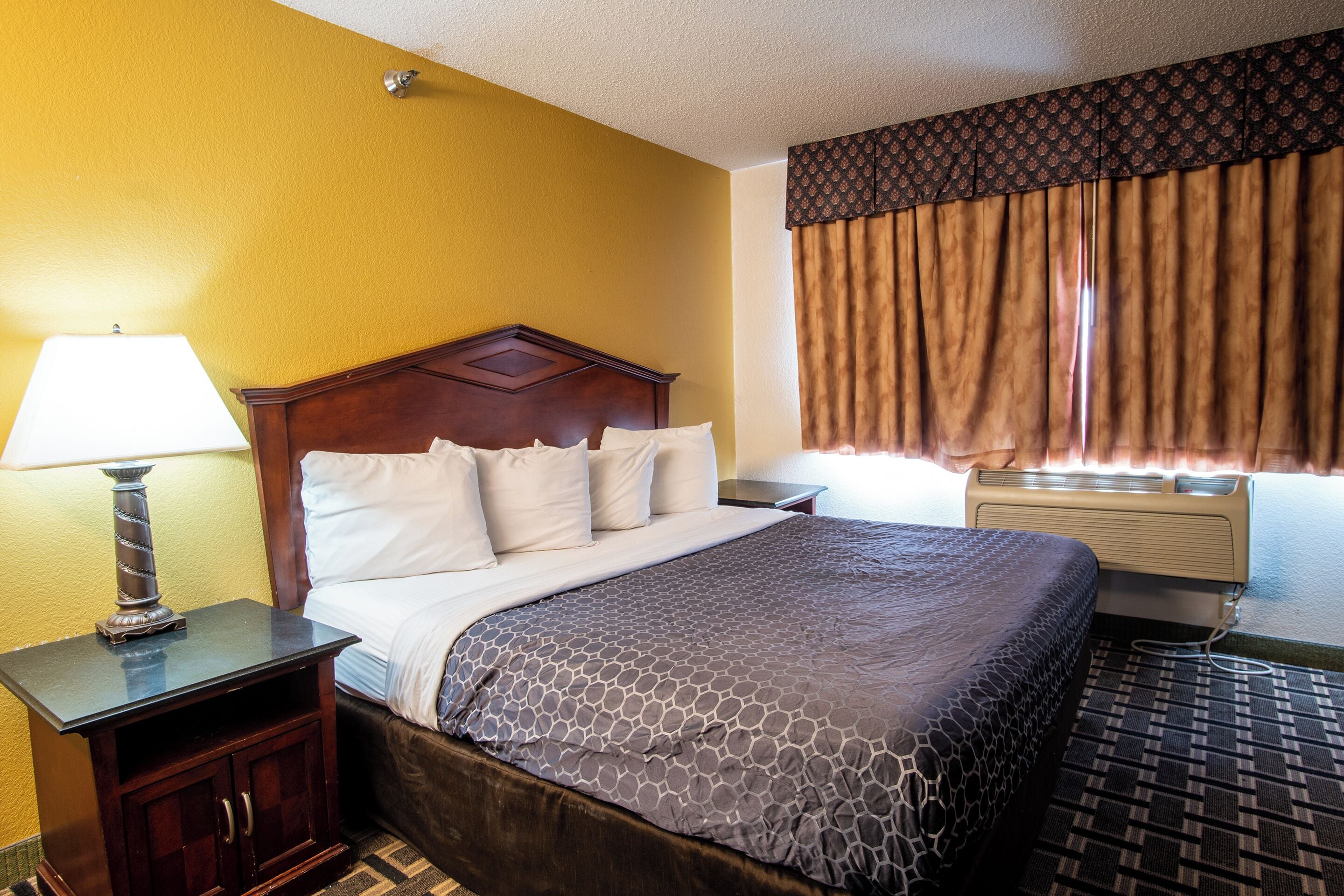 Une chambre d’hôtel avec un lit, une table de chevet avec une lampe, une commode et une fenêtre avec des rideaux.
