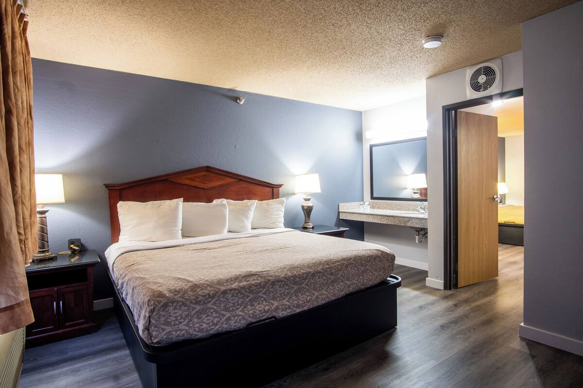 A hotel room with a large bed, bedside lamps, a wooden headboard, a nightstand, a mirror, and a door leading to another room.