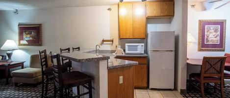 Cocina pequeña con armarios de madera, un microondas y un refrigerador. Hay taburetes de bar y un bote de basura.