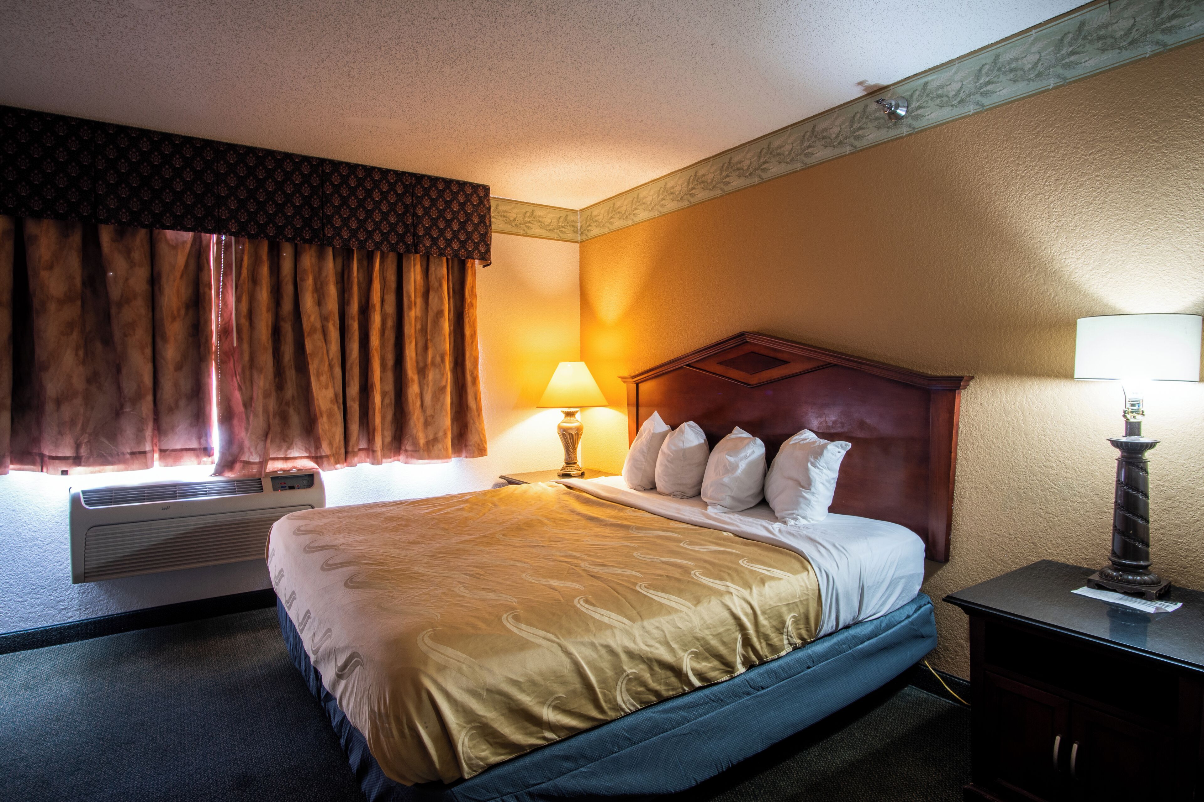 Quarto de hotel com uma cama grande, mesa de cabeceira com luminária, janela com cortinas e ar condicionado.