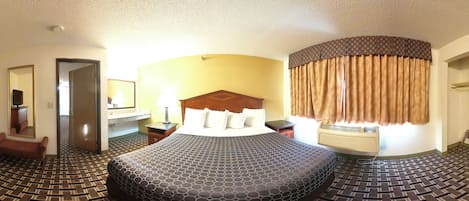 A hotel room with a large bed, a desk with a lamp, a chair, and a bathroom visible through an open door.