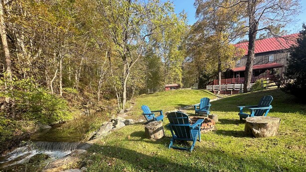 Un espacio al aire libre rústico con sillas Adirondack y un brasero, rodeado de un exuberante jardín y una pequeña cascada.