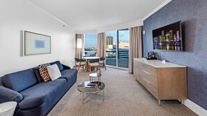 Ein modernes Hotelzimmer mit blauem Sofa, einem kleinen Couchtisch, einem an der Wand montierten Flachbildfernseher, einem Essbereich mit Tisch und Stühlen sowie Blick auf die Stadt durch große Fenster.