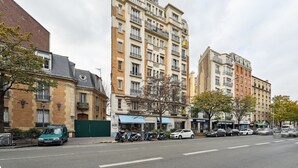 Un immeuble à plusieurs étages, avec une rangée de boutiques au rez-de-chaussée, des voitures garées et un arbre au premier plan.