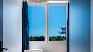Une pièce avec un plancher en bois, une chaise blanche, une fenêtre donnant sur un paysage verdoyant et un luminaire au plafond.