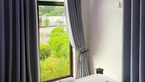 A hotel room with a bed, a window with curtains, an air conditioning unit, and a view of greenery outside. - Marbella lodge  (Bagamoyo)