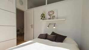 Ein Schlafzimmer mit einem Bett, zwei Kissen, einem Nachttisch, einer Wandablage mit Dekoration und einer Tür zu einem anderen Raum.