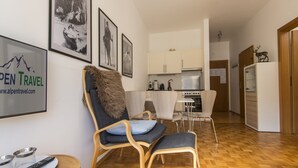 Ein modernes Wohnzimmer mit Holzboden, einer Couch mit Felldecke, einem Essbereich mit weißem Tisch und Stühlen sowie einer Küche mit weißen Schränken und einem Kühlschrank.
