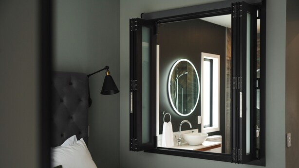 A bedroom with a bed, a bedside lamp, and a large mirror reflecting a bathroom with a sink and toilet. - The Residence Apartment Two (New Plymouth)