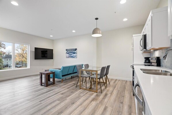 Una sala moderna con un sofá azul, una mesa de comedor y sillas. Se aprecia una zona de cocina con un lavaplatos y armarios.