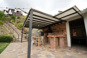 Un patio exterior con zona de barbacoa de ladrillo, un área de estar cubierta y una escalera que sube a una casa.