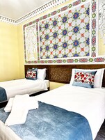 Habitación de hotel con dos camas, cada una con colcha azul y ropa blanca. La habitación tiene un tapiz grande y colorido con motivos geométricos sobre las camas.