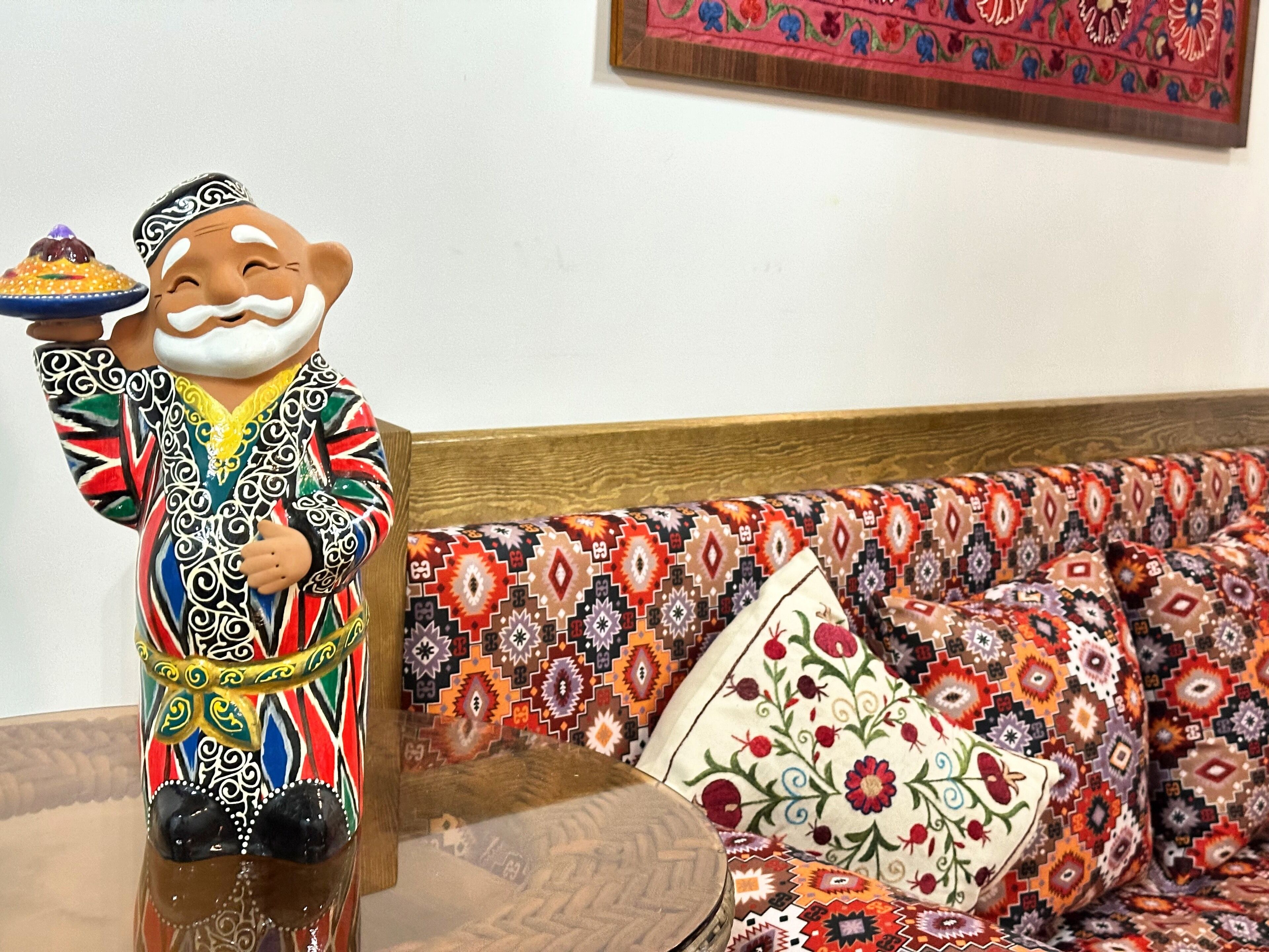 Una figura decorativa de cerámica con una bandeja, sobre una mesa de madera frente a un sofá estampado.