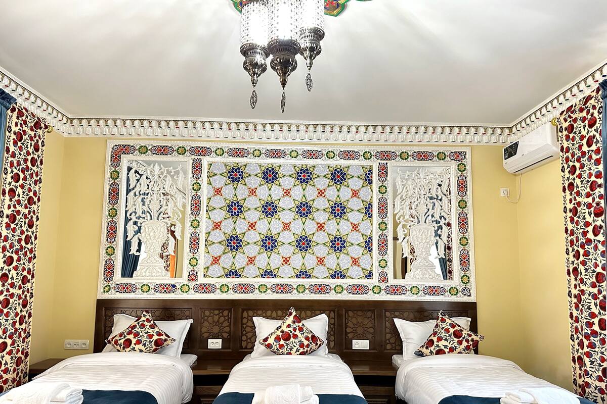 Quarto de hotel com três camas, um candelabro, azulejos decorativos e cortinas florais.