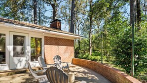 Un patio con dos sillas Adirondack y una mesa, un muro de ladrillo y un área techada con una puerta corrediza de vidrio.