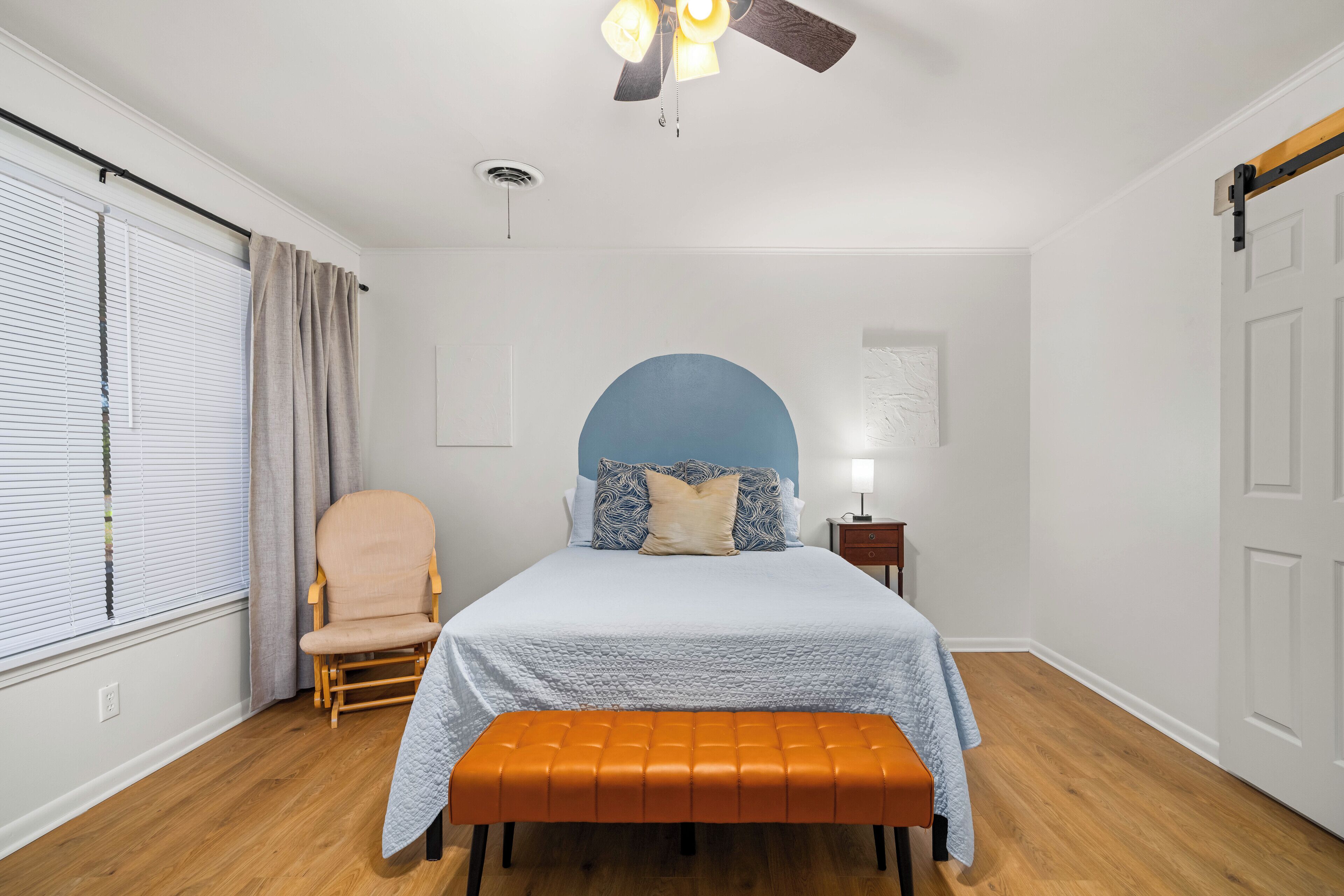 Een slaapkamer met een bed, een stoel, een bankje, een plafondventilator en een raam met gordijnen.