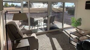 Una habitación iluminada por el sol con un ventanal corredero que da a un patio exterior. En el interior, hay un sofá, una mesita y una silla.