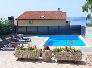 A swimming pool with a view of the sea, a covered patio with lounge chairs, and stone planters with flowers. - Spacious Novi Vinodolski Apartments With Terrace and Shared Pool (Novi Vinodolski)