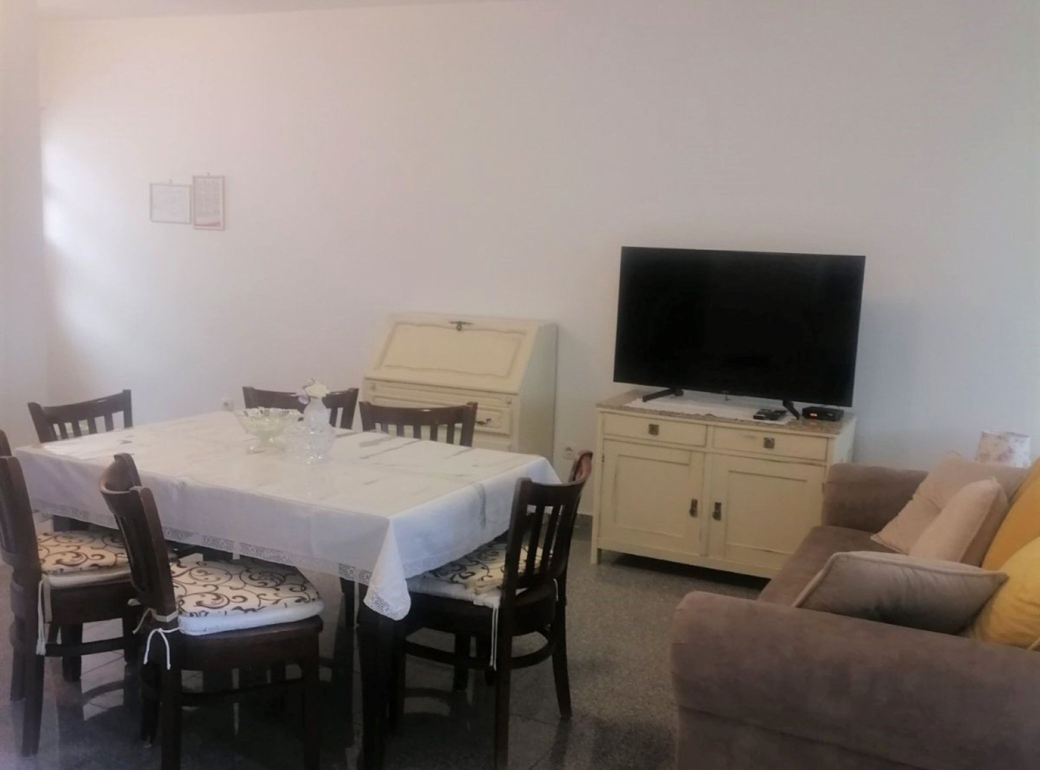 Un comedor con una mesa dispuesta para cuatro personas, un televisor sobre un mueble bajo y un sofá al fondo.