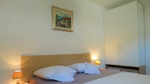 A hotel room with a wooden headboard, two white pillows, a bed with a white duvet, a nightstand with a lamp, a wardrobe, and a painting on the wall. - Spacious Novi Vinodolski Apartments With Terrace and Shared Pool (Novi Vinodolski)