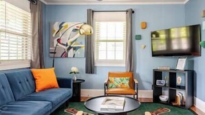 Un salon avec un canapé bleu, une table basse ronde, un fauteuil coloré, un téléviseur et une bibliothèque.