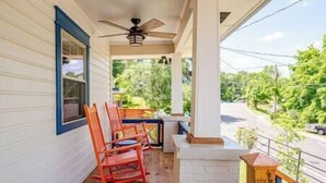 Une véranda dotée d’un ventilateur de plafond, d’une chaise berçante en bois et offrant une vue sur une route et la verdure environnante.
