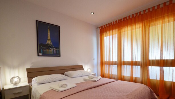 A hotel room with a bed, a nightstand, a lamp, a painting of the Eiffel Tower, and orange curtains. - Sleek and Serene Flat and Rooms - Studio Apartment with Terrace (Rovinj)