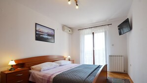 Ein Schlafzimmer mit einem hölzernen Bett, Nachttischlampen, einem an der Wand montierten Fernseher, einem Fenster mit durchsichtigen Vorhängen und einem Heizkörper.