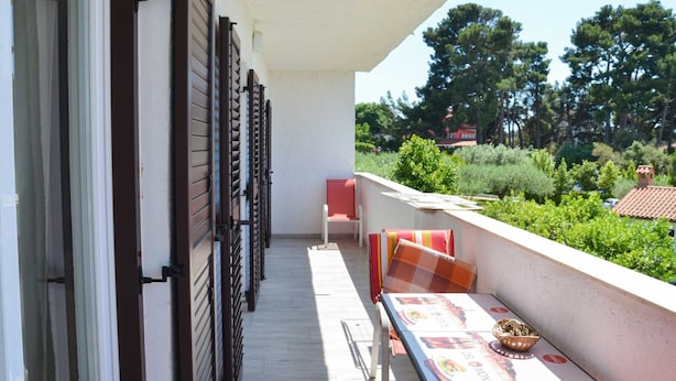 Un balcón con una mesa y sillas, vistas a árboles y edificios, y una puerta con contraventanas.