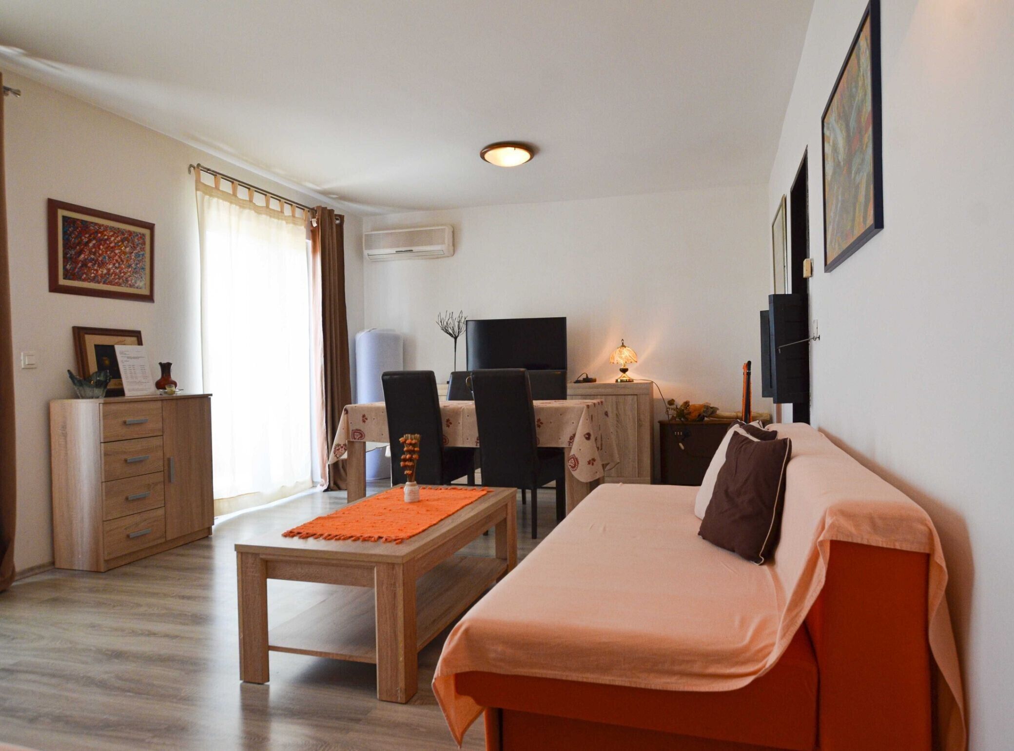 A cozy room with a bed, a desk, and a chair. There's a window with curtains, a wall-mounted air conditioner, and a framed picture.