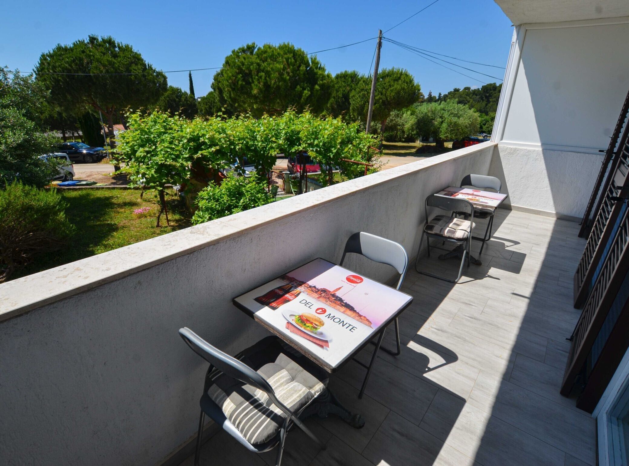 Un balcon avec deux tables et des chaises, une vue sur un jardin avec des arbres et une voiture, et un bâtiment à l’extérieur blanc.