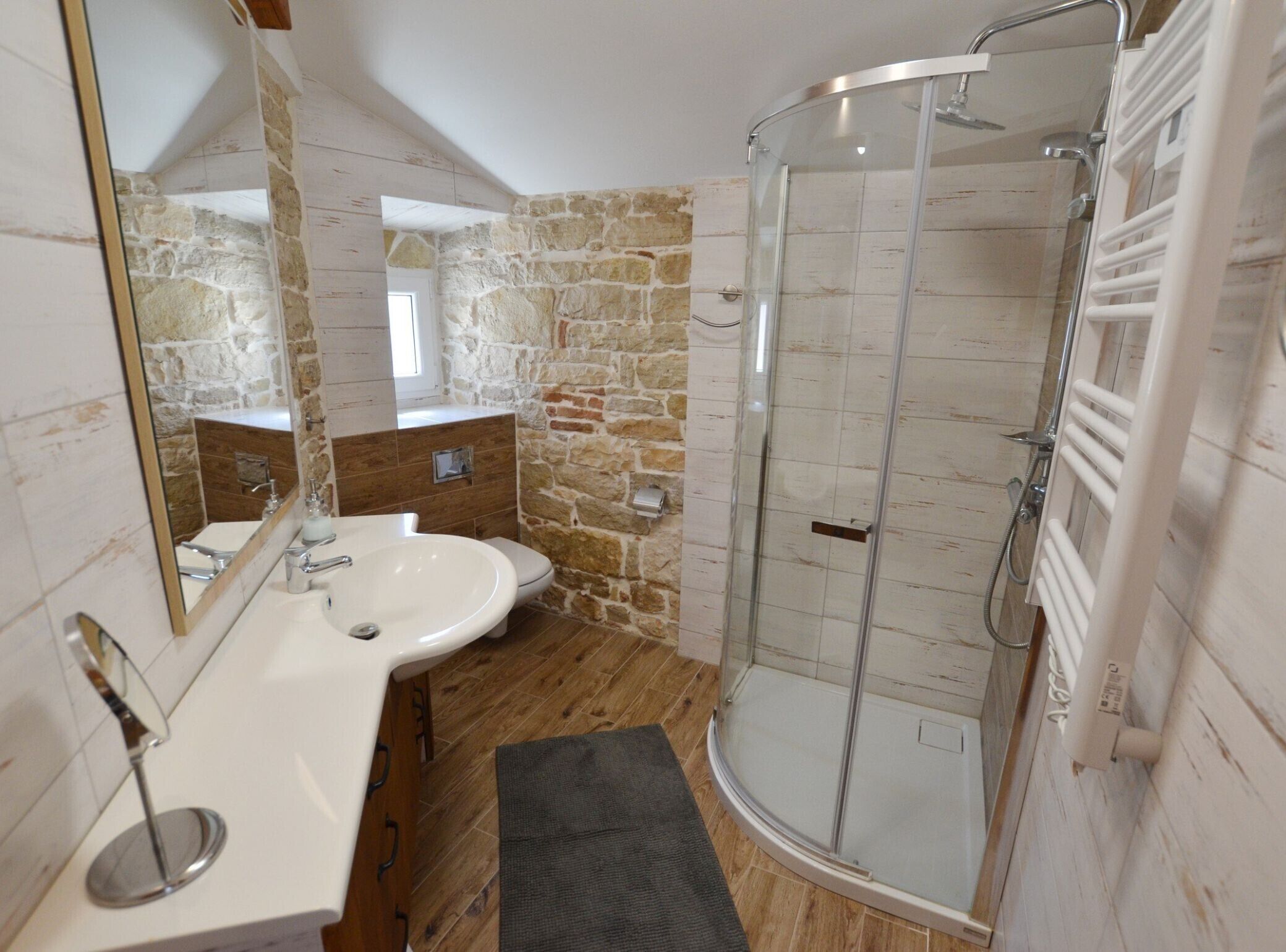 Une salle de bain avec un mur en pierre, une douche en verre, un lavabo blanc et un miroir.
