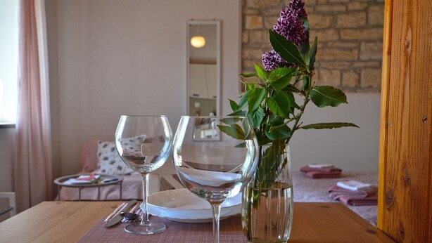 Een eettafel gedekt met witte borden, twee wijnglazen en een vaas met een paarse bloem.