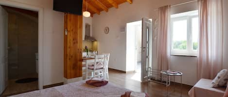 Un dormitorio con cama, mesita de noche, silla, pequeña mesa, ventana con cortinas y techo de madera.