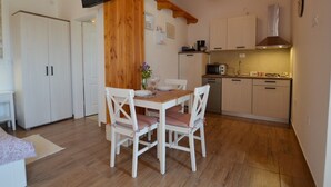 Een kleine keuken met houten kastjes, een eettafel met stoelen, een televisie aan de muur en een klok.