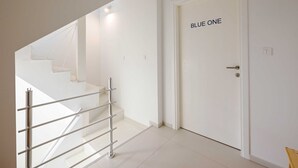 Un intérieur moderne avec un escalier, une porte blanche portant l’inscription « BLUE ONE », et une rambarde en métal.
