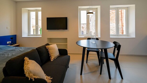 Un salon moderne comprenant un canapé, une table à manger avec des chaises, un téléviseur fixé au mur et de grandes fenêtres laissant entrer la lumière naturelle.