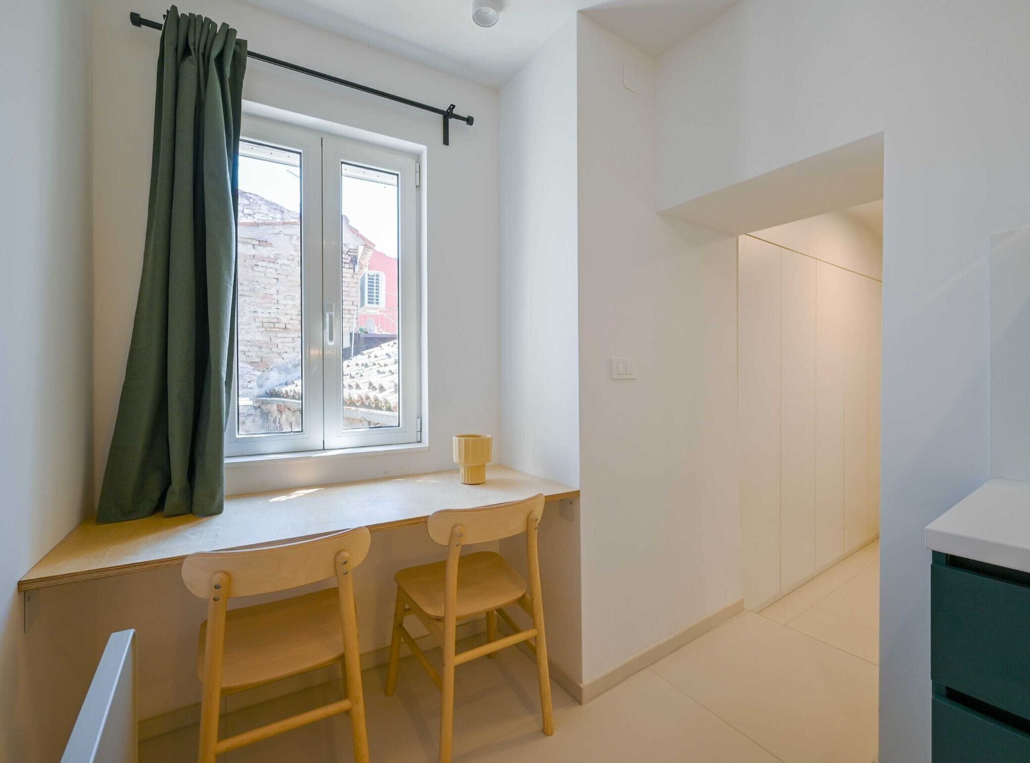 A small, bright room with a window, a desk, two wooden chairs, and a white cabinet.