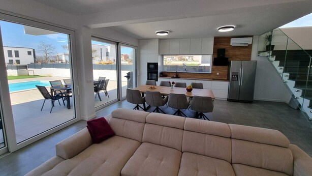 Ein modernes Wohnzimmer mit einem beigefarbenen Sofa, einem Essbereich mit Holztisch und -stühlen, einer Küche mit weißen Schränken und einem eingebauten Backofen sowie einer Glasstiege.