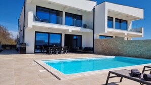 Ein modernes, zweistöckiges Haus mit Pool, Sitzgelegenheiten im Freien und einer Steinmauer.