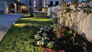 Un jardin bien entretenu, agrémenté de diverses fleurs et doté d’une pelouse soigneusement taillée, se trouve devant un bâtiment en pierre, orné de nombreuses fenêtres et d’un balcon.