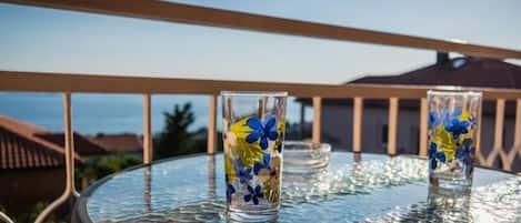 A round glass table with two clear glasses featuring blue and yellow floral patterns, set against a balcony railing with a clear sky in the background.