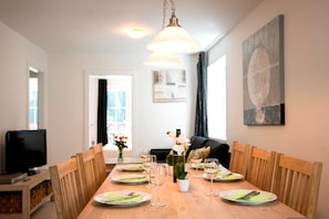 Un coin repas avec une table en bois dressée pour six personnes, entourée de chaises en bois. On aperçoit un téléviseur au fond, et un tableau est accroché au mur.